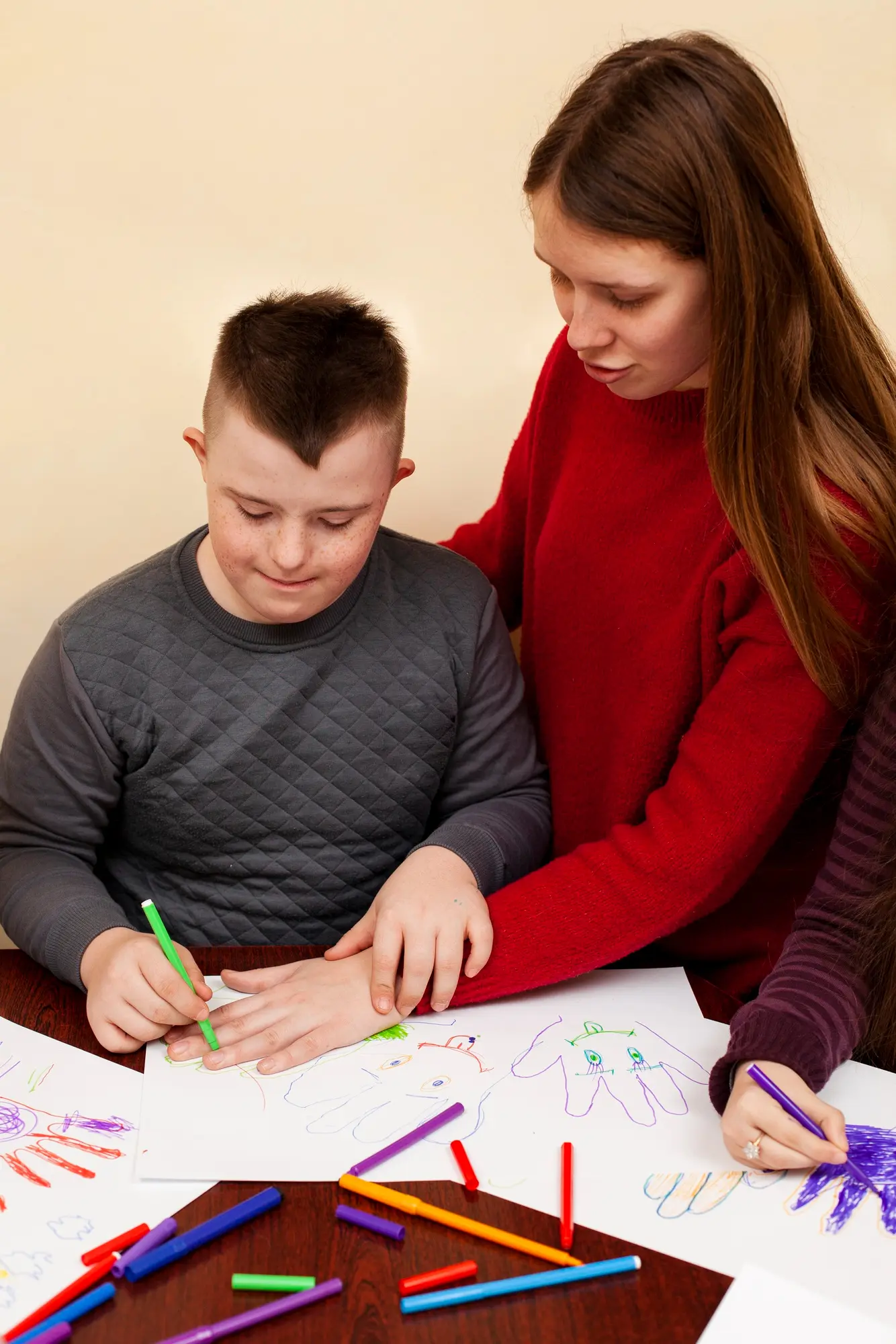A boy and a volunteer draw together at a table covered with colored markers.