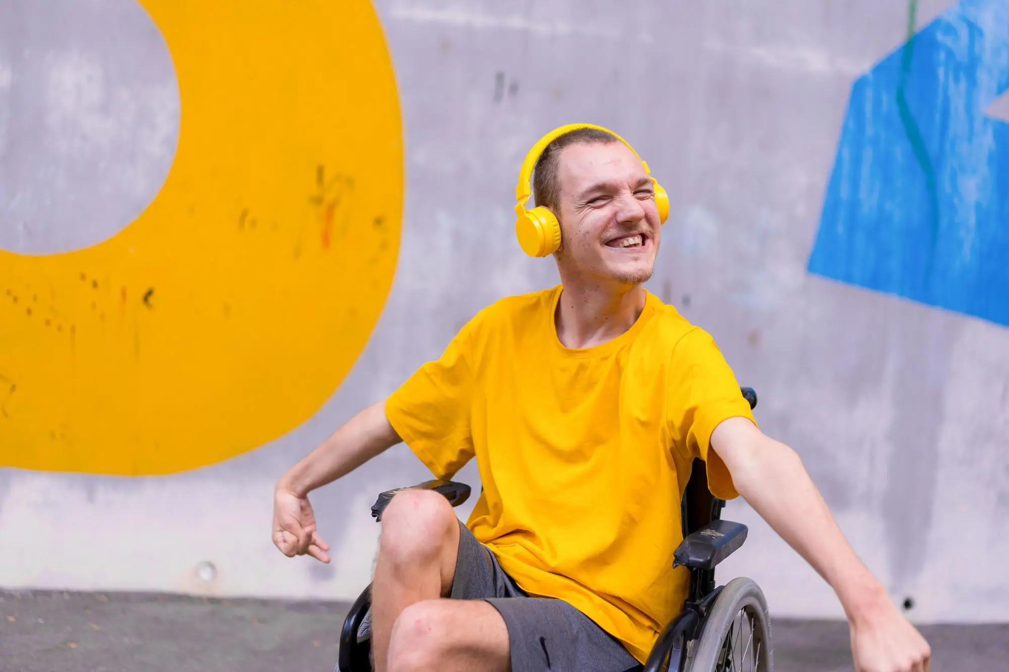 A young man in a yellow shirt and headphones laughs joyfully in his wheelchair in front of a bright painted wall.